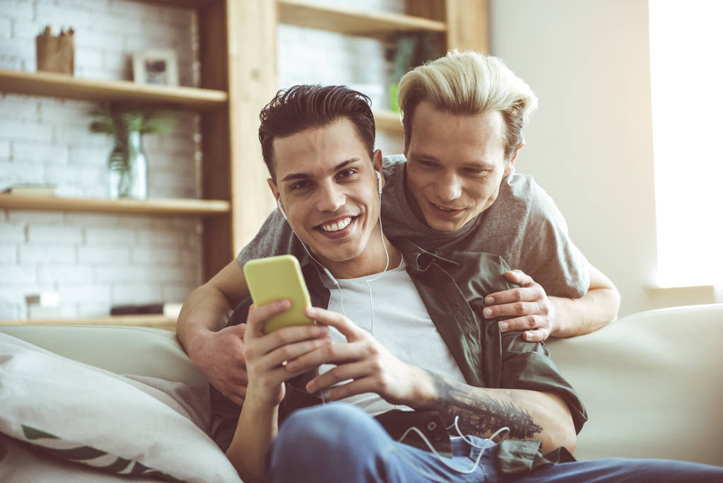 Jovem casal gay usando smartphone e abraçando em casa
 - Foto, Imagem