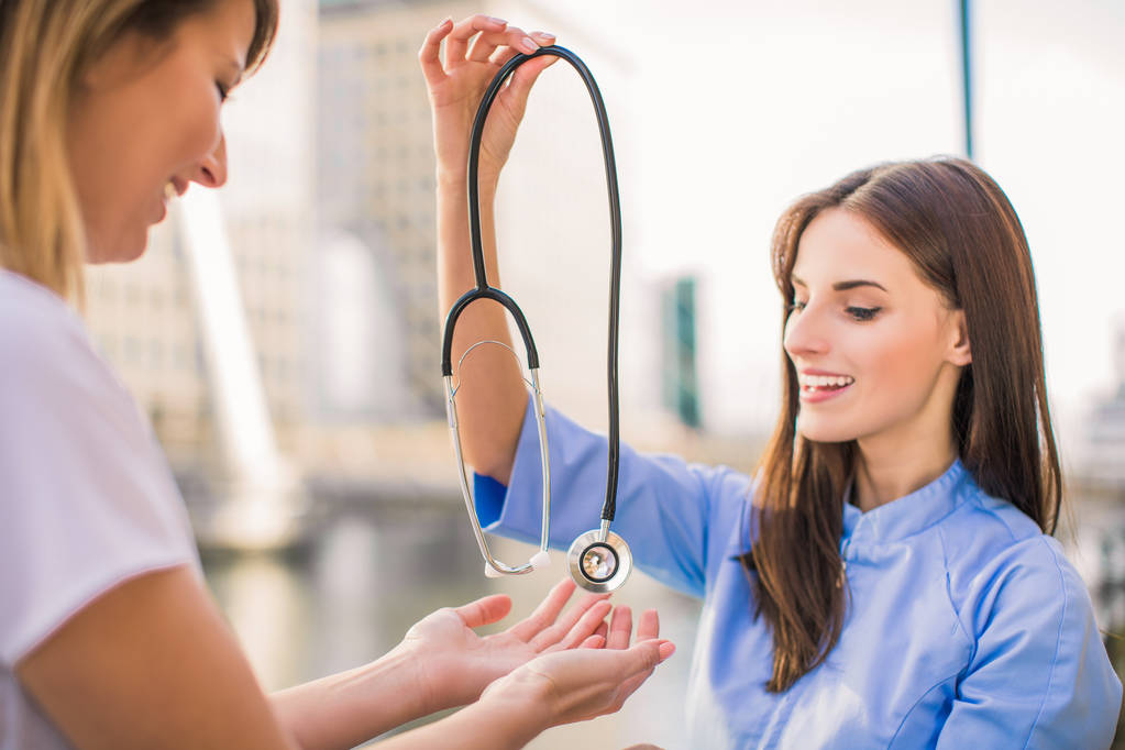 Close-up lachende jonge verpleegster geeft stethoscoop tot vrouwelijke arts, buiten - Foto, afbeelding