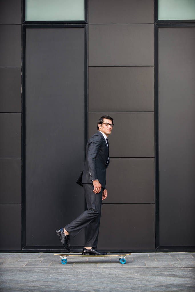 Homem bonito jovem com terno de negócios montando em um longboard - retrato empresário corporativo, conceitos sobre negócios, mobilidade e estilo de vida
 - Foto, Imagem