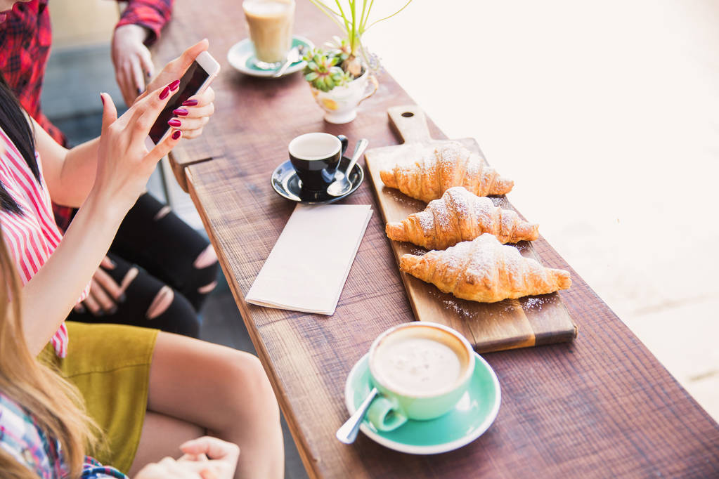 As pessoas sentam-se à mesa com os croissants dourados e xícaras de café, um deles mantém o telefone, no café.
 - Foto, Imagem