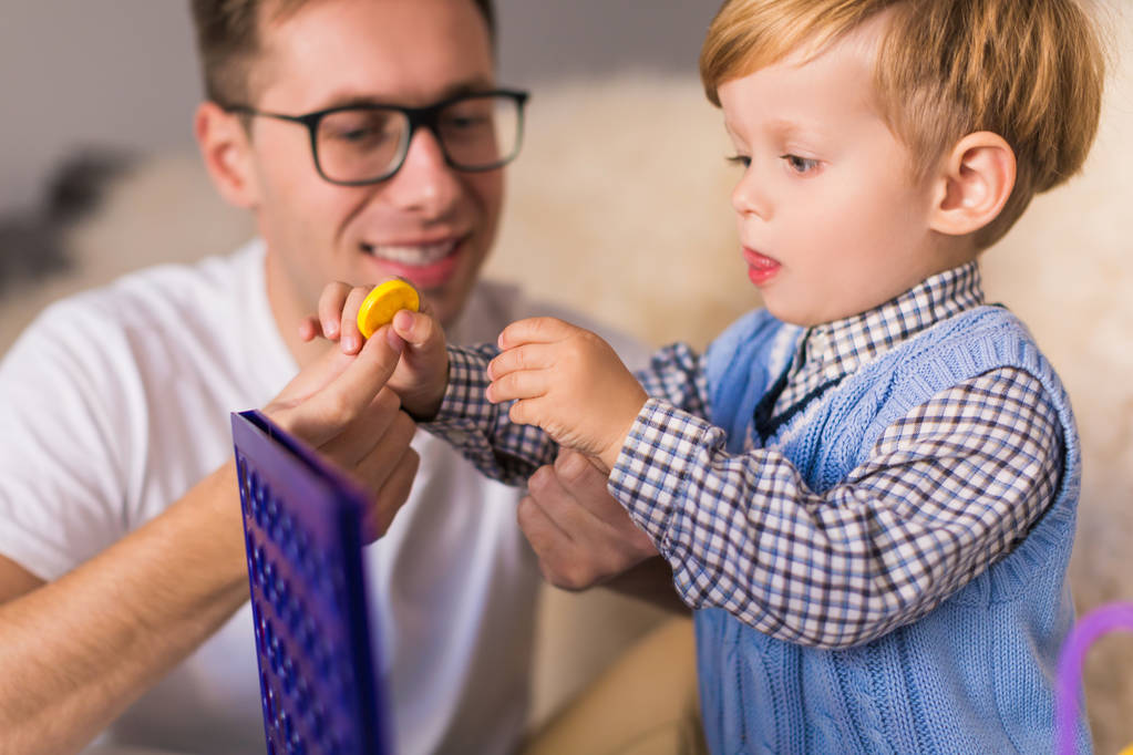 Porträt eines kleinen niedlichen, ernsthaften Sohnes, der in der Nähe seines lächelnden Vaters zu Hause mit Entwicklungsspielzeug spielt - Foto, Bild