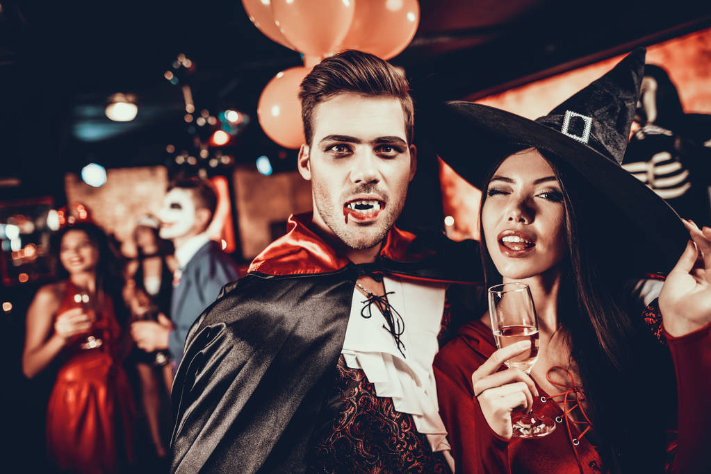 Молодая счастливая пара в костюмах на Хэллоуинской вечеринке. Красивый мужчина и красивая женщина пьют шампанское на Хэллоуинской вечеринке в ночном клубе. Друзья веселятся. Празднование Хэллоуина
 - Фото, изображение