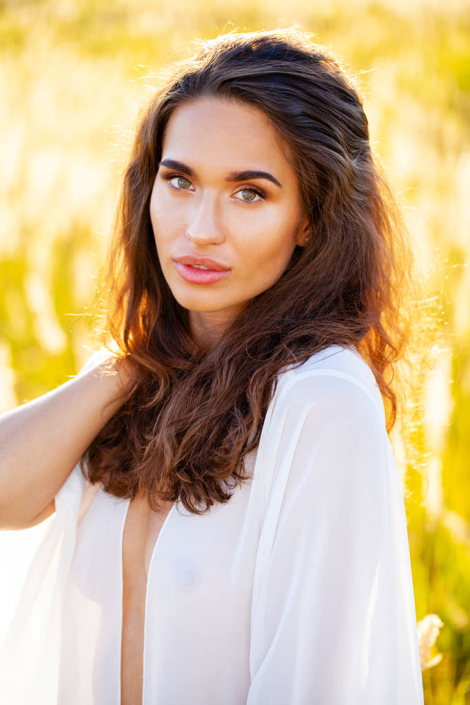 Portrait close up of young beautiful brunette woman in white dress, posing at sunset in the field - Photo, Image