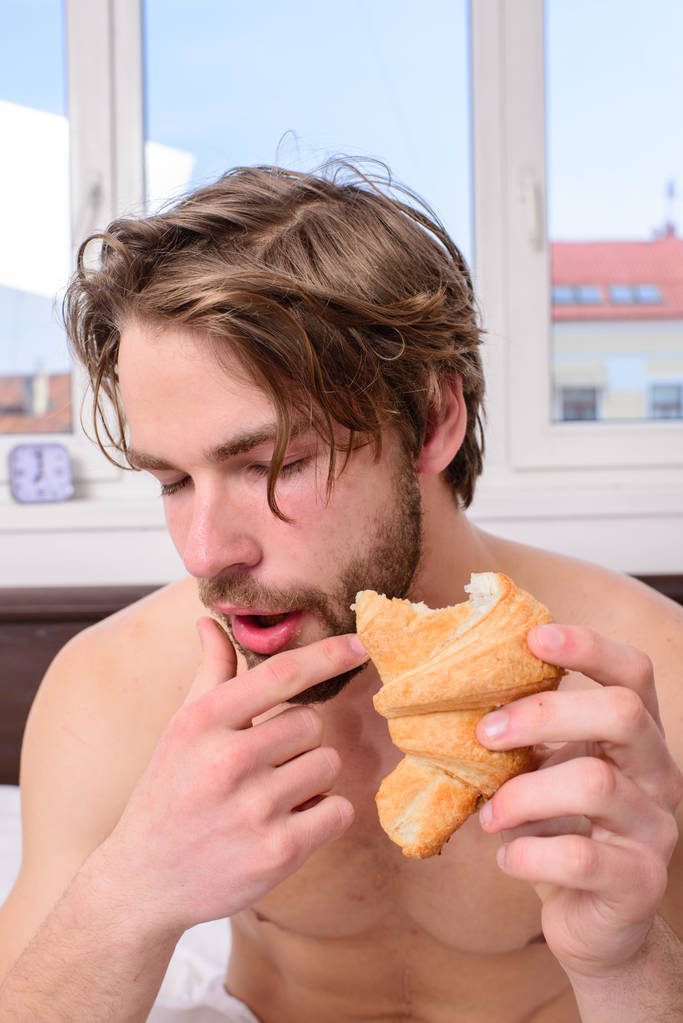 Mies syö croissantia hän pitää leipomotuotteista. Kaveri puree makeaa jälkiruokaa. Mies parrakas komea kaveri syö aamiaista sängyssä. Kaveri pitää croissant makuuhuone hotellihuoneen ikkuna tausta. Nauti jokaisesta puraisusta
 - Valokuva, kuva