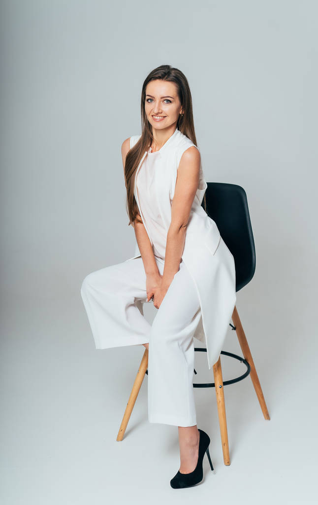 a pretty business woman sits on a chair and smiles on a white studio background. Young business lady is dressed in a stylish white suit with a long jacket. Close-up - Photo, Image