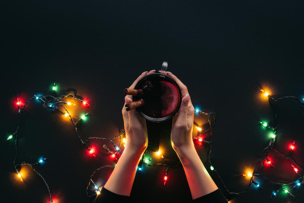 黒のクリスマス コンセプト上分離されたガーランド近くグリュー ワインのカップを保持している女性のトリミングされた画像 - 写真・画像