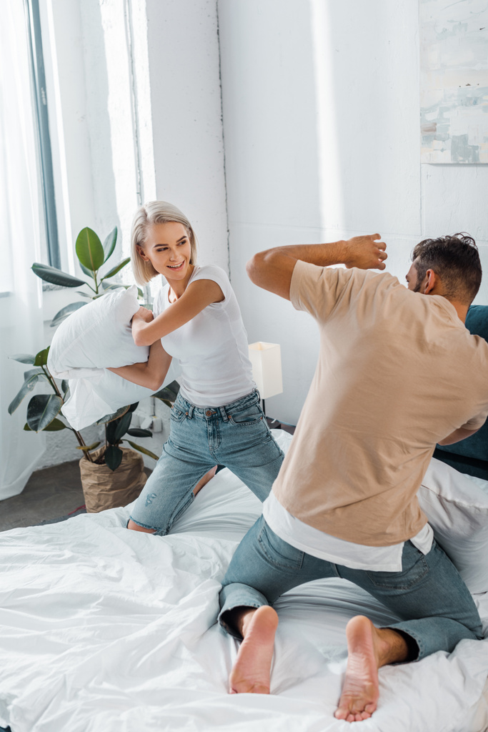 happy young couple fighting with pillows on bed in bedroom - Photo, Image