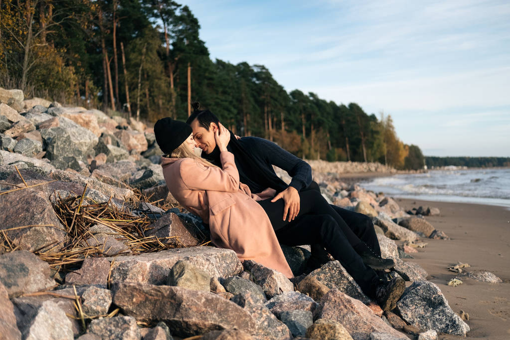 Young sexual couple in love lying down on stones at the beach, hugging kissing and enjoy each other. Affectionate and passionate elegant couple seducing and flirting. Autumn weather, seaside coastline - Photo, Image