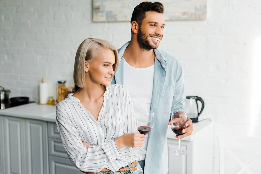 Lächelndes junges Paar hält Rotweingläser in der Küche und schaut weg - Foto, Bild