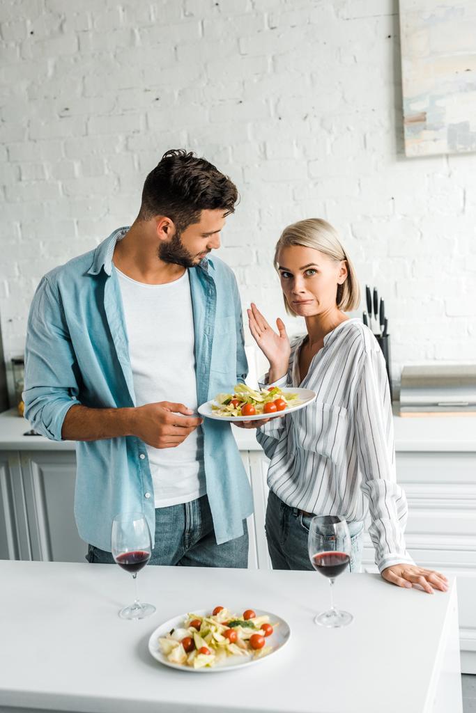 geïrriteerde vriendin grimassen en gebaren in de buurt van vriendje met plaat van salade in keuken - Foto, afbeelding