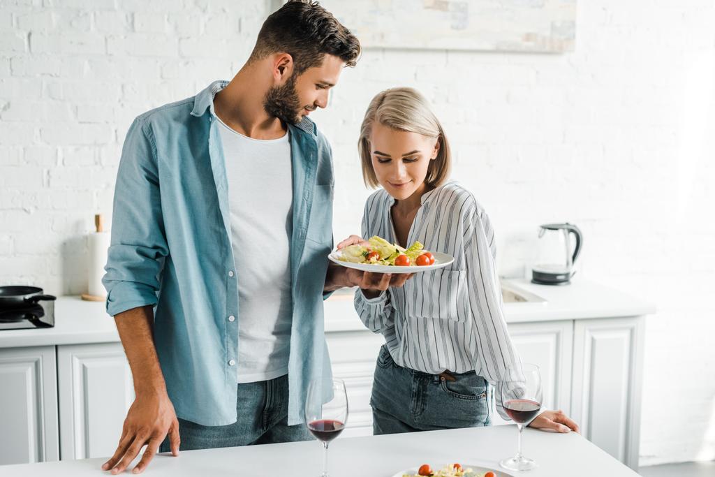 парень показывает салат на тарелке привлекательной девушке на кухне
 - Фото, изображение