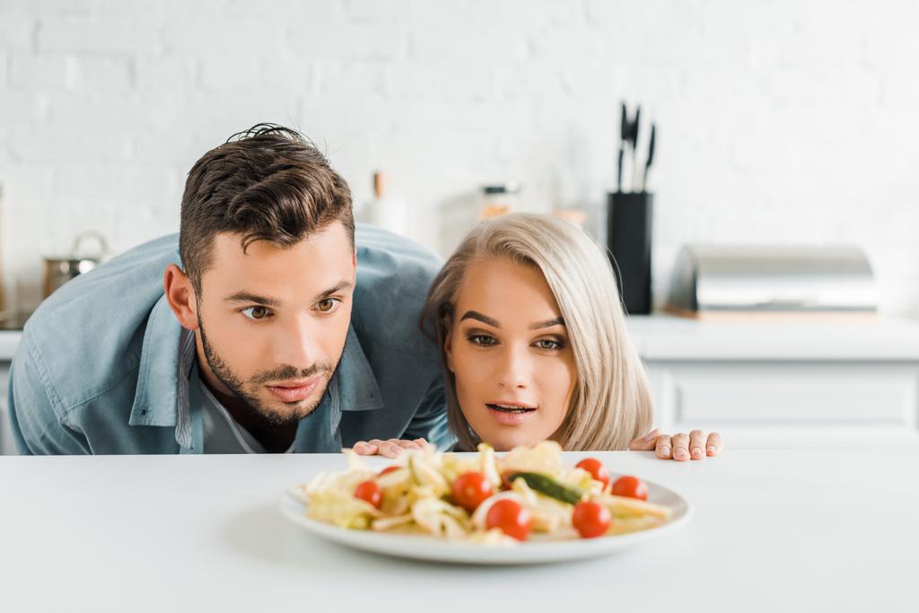 surprised girlfriend and boyfriend looking out from table and looking at plate with salad in kitchen - Photo, Image