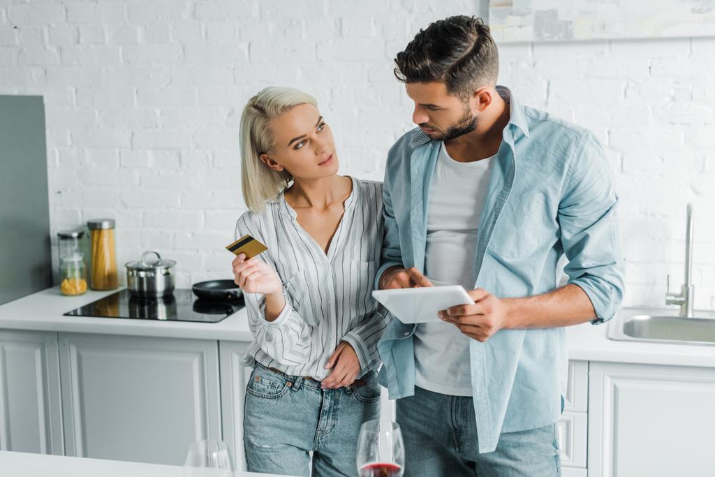 namorado segurando tablet e namorada de pé com cartão de crédito na cozinha
 - Foto, Imagem