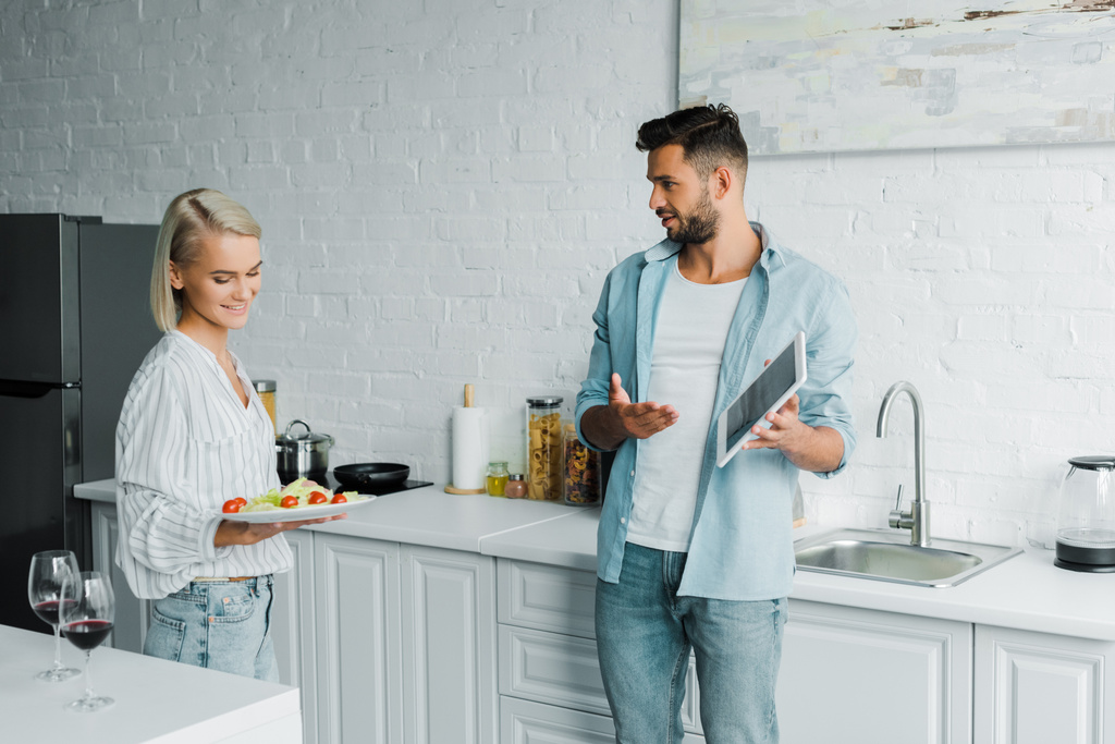 boyfriend pointing on tablet to smiling girlfriend with dish in kitchen - Photo, Image