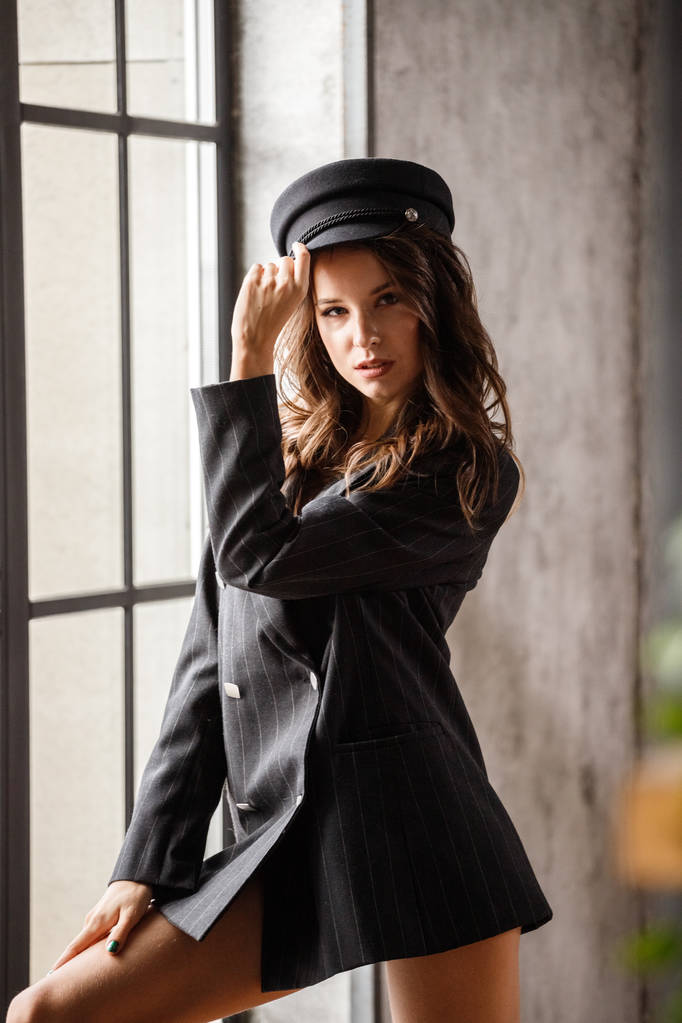 Güzellik kadın modeli ayakta pencere yakın portresi. Modern iç gri ceket ve gri şapka giymiş esmer kadın - Fotoğraf, Görsel