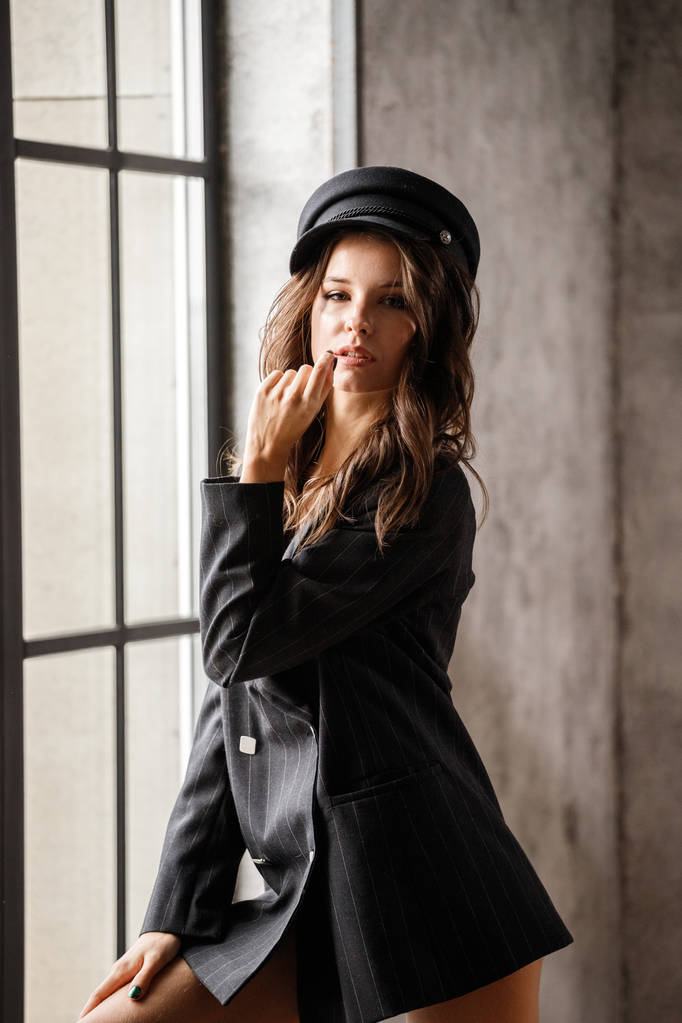 Портрет женщины-модели, стоящей у окна. Брюнетка в серой куртке и серой шляпе в современном интерьере
 - Фото, изображение