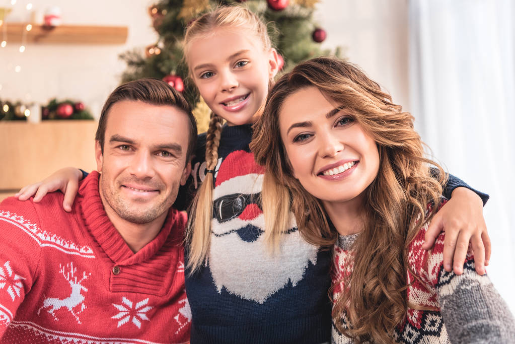 усміхнені батьки і дочка в зимових светрах обіймаються і дивляться на камеру вдома
 - Фото, зображення