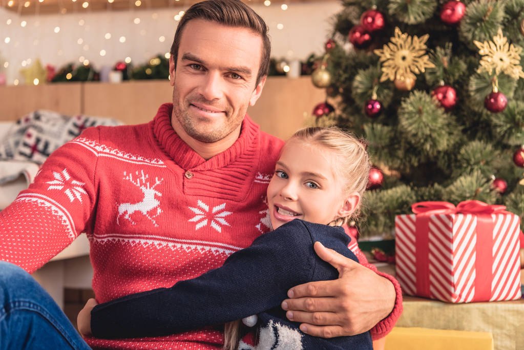 Отец обнимает очаровательную дочь возле рождественской елки дома, и они смотрят в камеру
 - Фото, изображение