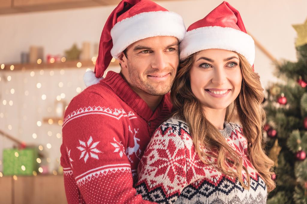 自宅のクリスマス ツリーと離れている彼らの近くサンタ帽子ハグ妻に笑顔の夫 - 写真・画像