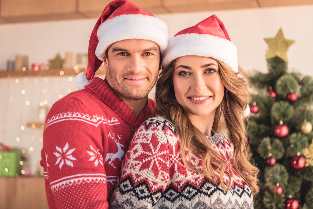 портрет мужа и жены в шляпах Санты, обнимающихся у рождественской елки дома и смотрящих в камеру
 - Фото, изображение