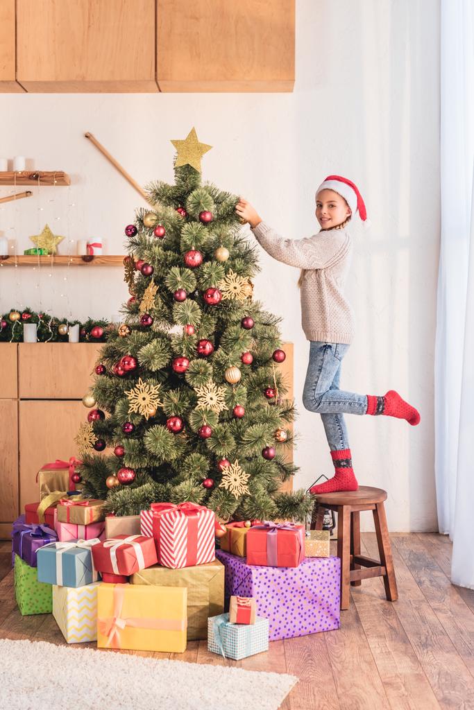 Lächelndes Kind mit Weihnachtsmütze, das auf einem Hocker steht und zu Hause den Weihnachtsbaum mit Geschenken schmückt - Foto, Bild
