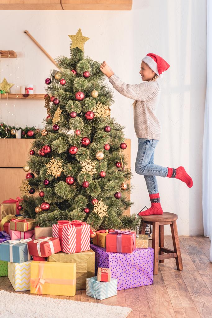 happy child in santa hat standing on stool and decorating christmas tree with gift boxes - Photo, Image