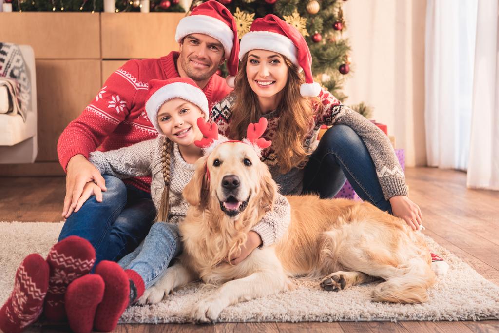 gelukkige familie in santa hoeden met hond in herten hoorns zitten in de buurt van de kerstboom - Foto, afbeelding