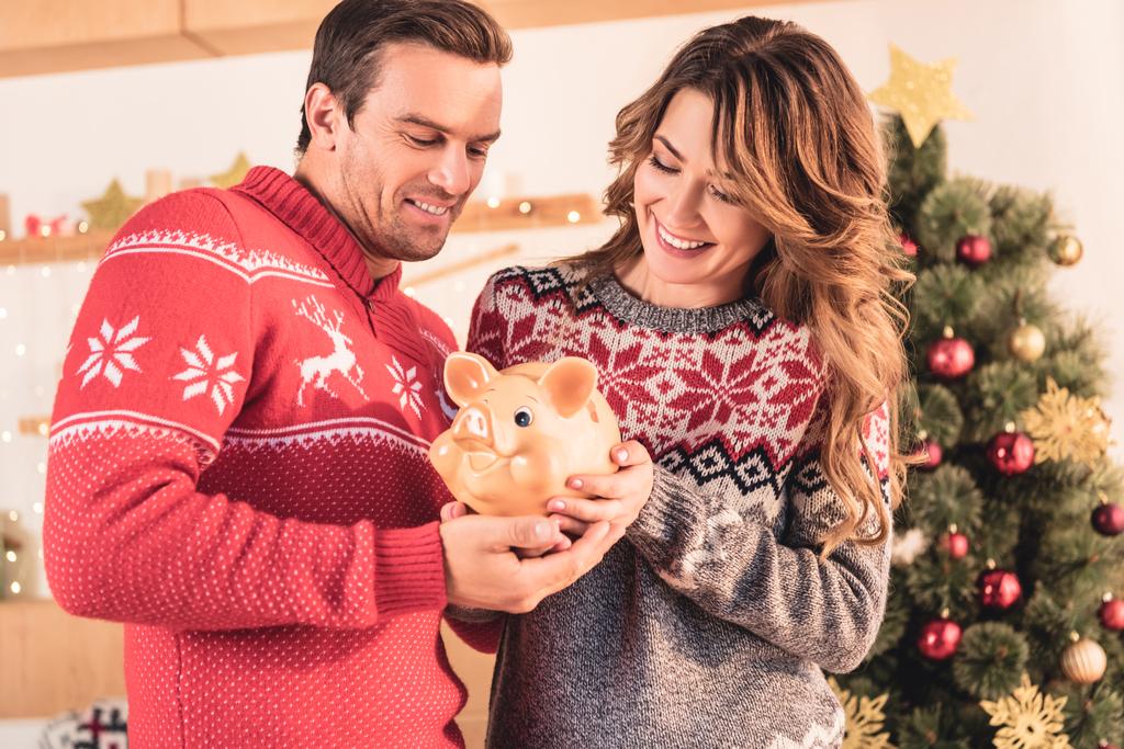χαμογελώντας ζευγάρι εκμετάλλευση κουμπαράς με εξοικονόμηση στο σπίτι με το χριστουγεννιάτικο δέντρο - Φωτογραφία, εικόνα
