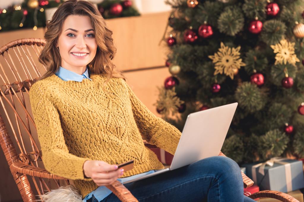 mujer sonriente de compras en línea con tarjeta de crédito y portátil en casa con árbol de Navidad
 - Foto, imagen