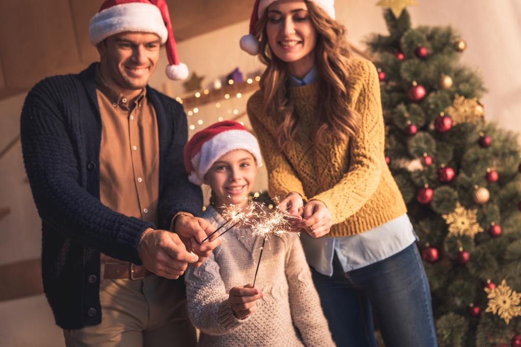 счастливая семья с дочерью в шляпах Санты держа искры дома с рождественской елкой
 - Фото, изображение