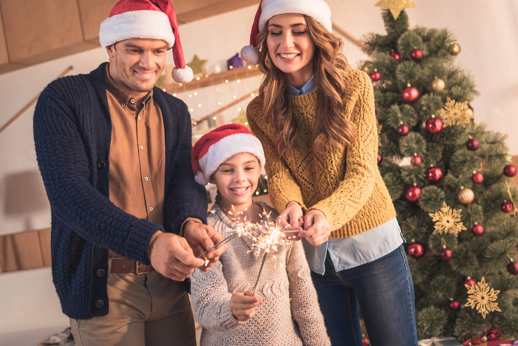 glückliche Mutter, Vater und Tochter in Weihnachtsmützen mit Wunderkerzen zu Hause mit Weihnachtsbaum - Foto, Bild