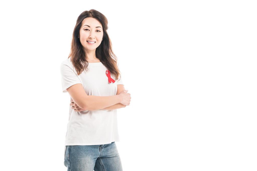szczęśliwy dorosły asian kobieta z aids świadomości czerwoną wstążką na t-shirt, patrząc na kamery z skrzyżowanymi rękami na białym tle - Zdjęcie, obraz