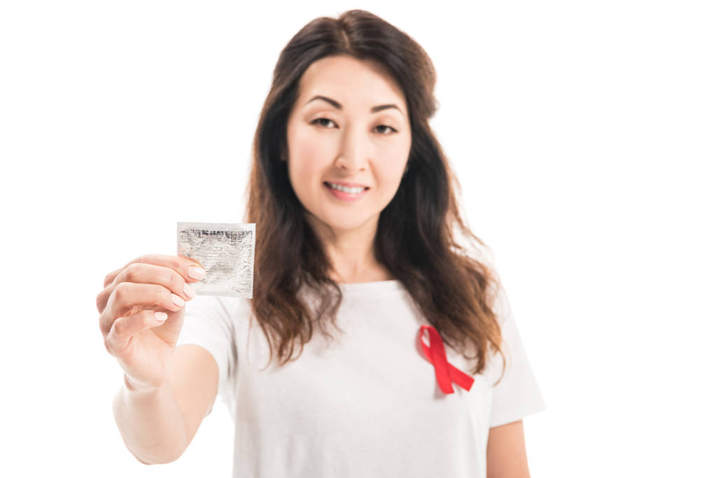 smiling adult asian woman with aids awareness red ribbon on t-shirt holding condom isolated on white - Photo, Image