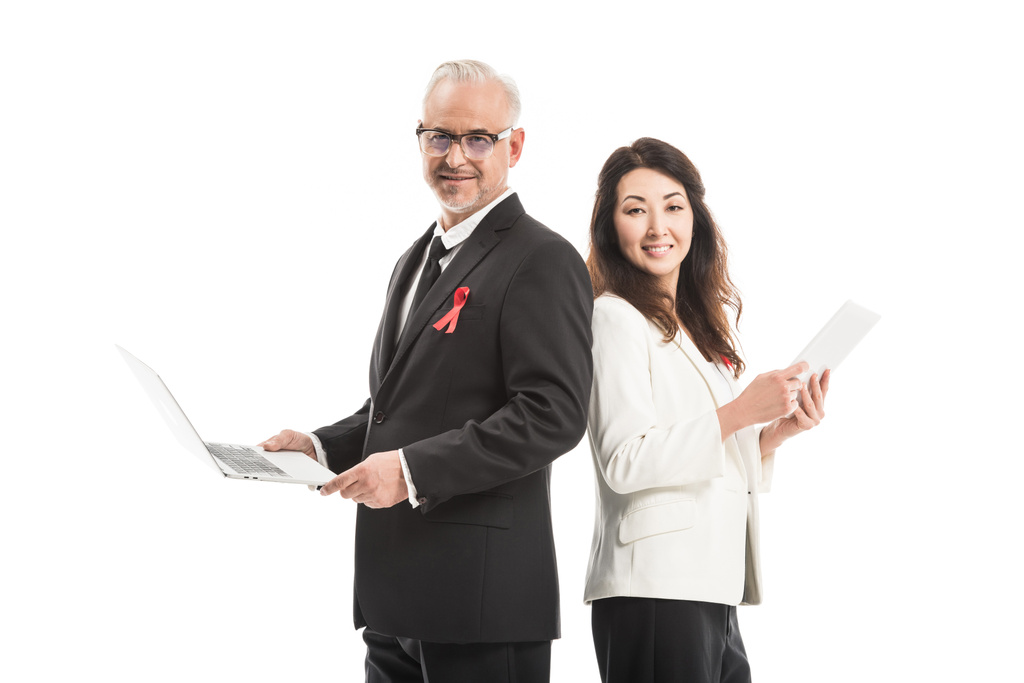 smiling adult businesspeople with aids awareness red ribbons working with gadgets while standing back to back and looking at camera isolated on white - Photo, Image