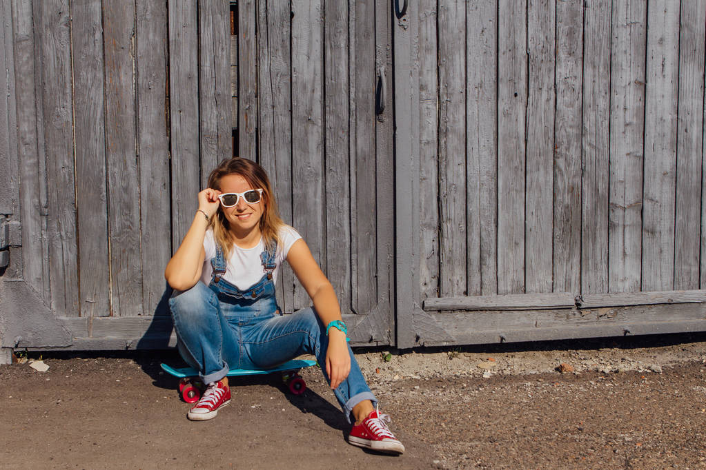 Porträt einer lächelnden Frau in Overalls und Sonnenbrille, die auf ihrem Skateboard neben der alten Holzwand sitzt. - Foto, Bild