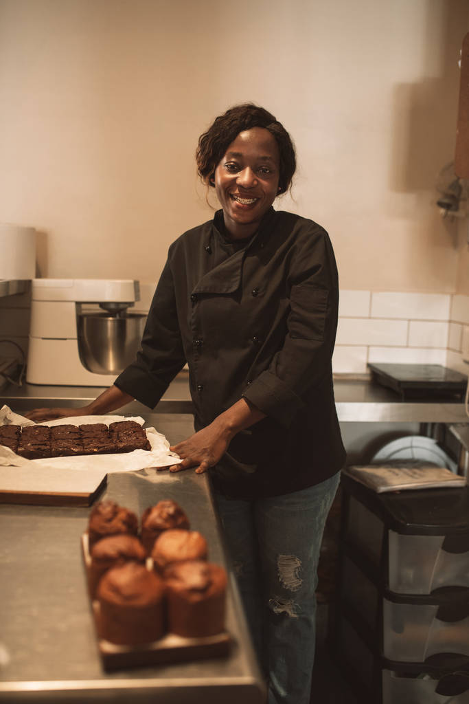 Портрет усміхнений африканських жіночий Бейкер, роблячи смачні тістечка під час роботи над лічильник в комерційний кухня  - Фото, зображення