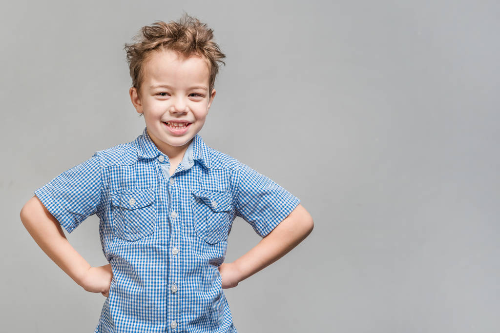 Симпатичный маленький мальчик в голубой рубашке стоял с руками на бедрах на сером фоне. Isolated
 - Фото, изображение
