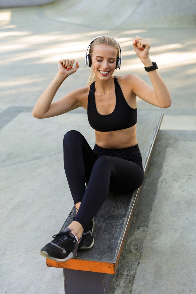 Изображение счастливой молодой фитнес-женщины в парке слушающей музыку в наушниках
. - Фото, изображение