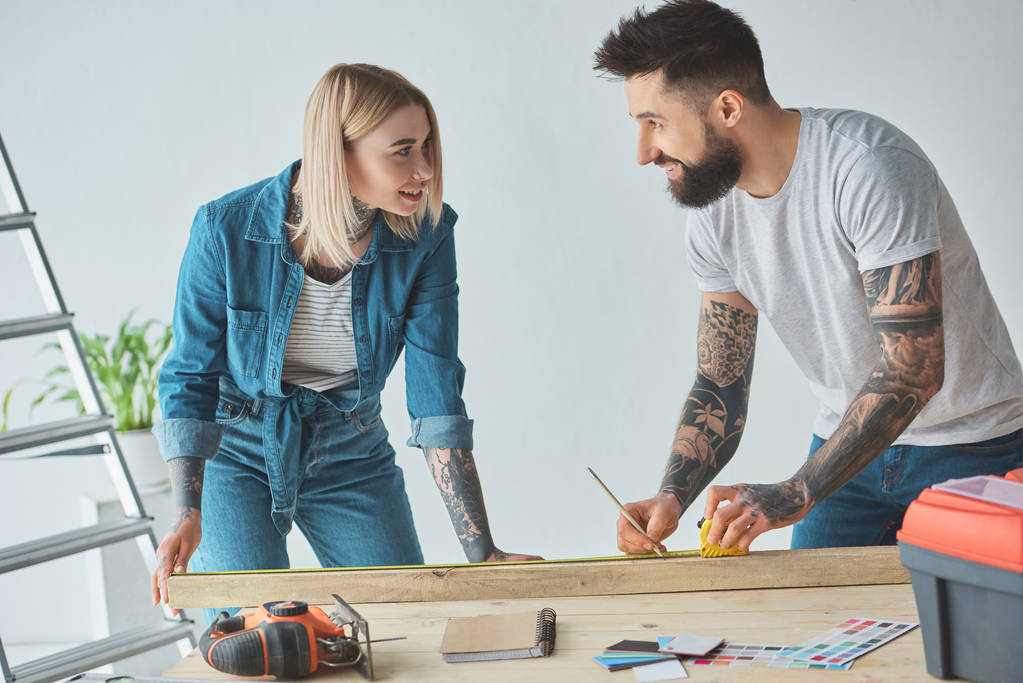 young tattooed couple smiling each other while holding pencil, wooden plank and measuring tape - Photo, Image