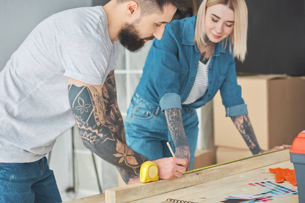 smiling young tattooed couple working with wooden plank and measuring tape  - Photo, Image