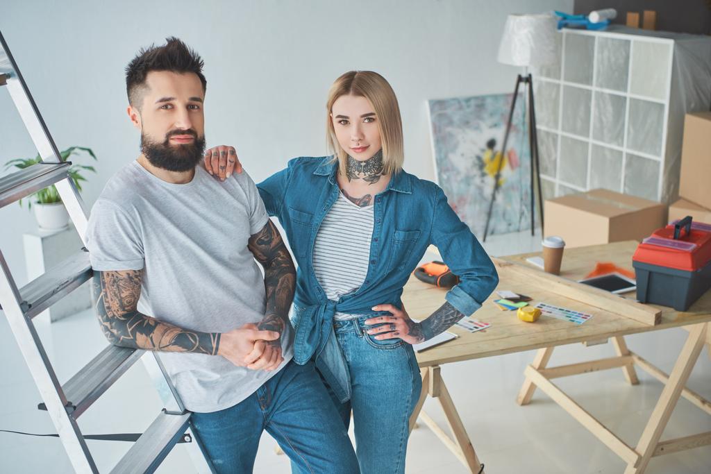όμορφο νεαρό ζευγάρι τατουάζ βλέπουν κάμερα ενώ στέκεται μαζί σε νέο σπίτι - Φωτογραφία, εικόνα