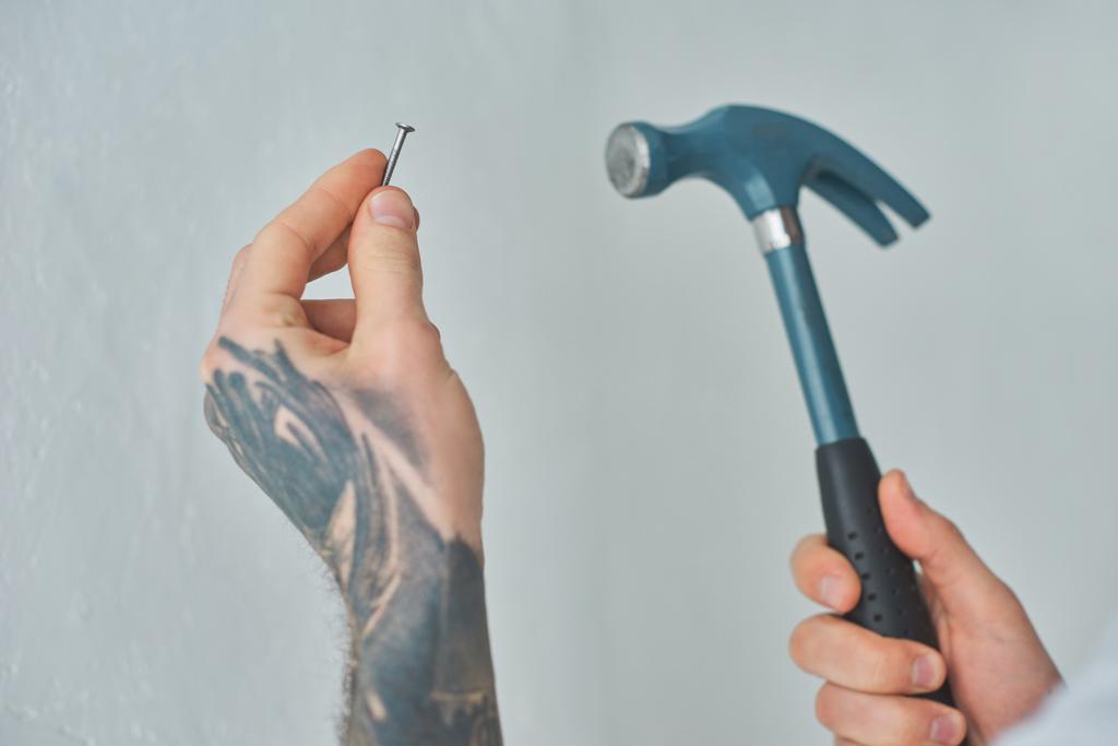 primer plano vista parcial de hombre joven tatuado sosteniendo martillo y clavo
 - Foto, Imagen