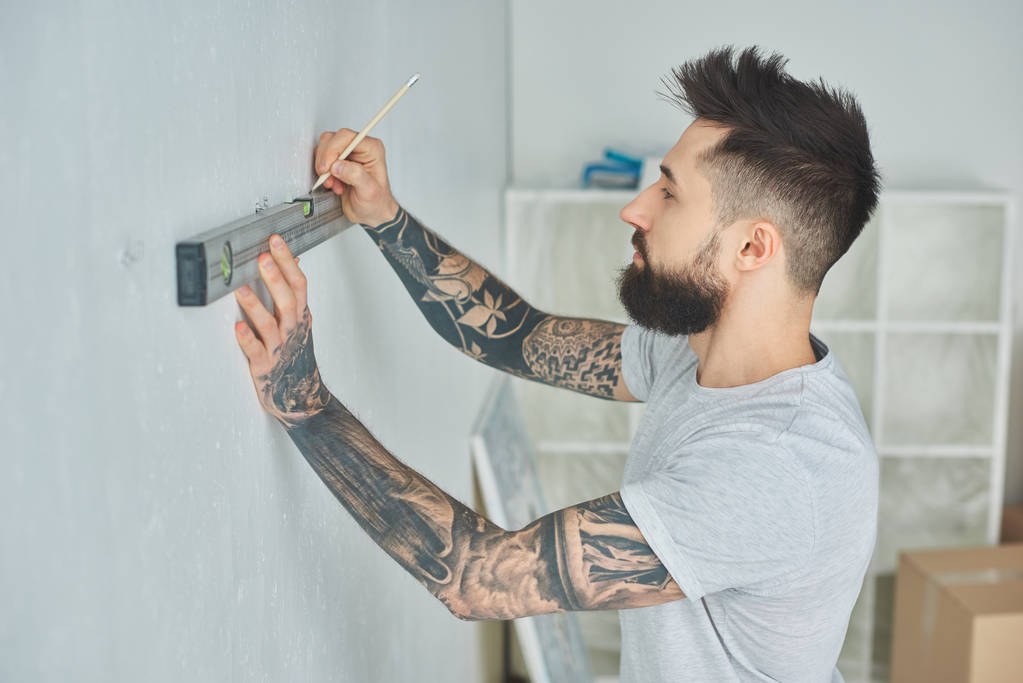 vue latérale du jeune homme tatoué tenant l'outil de niveau et le mur de marquage avec crayon pendant les réparations
 - Photo, image
