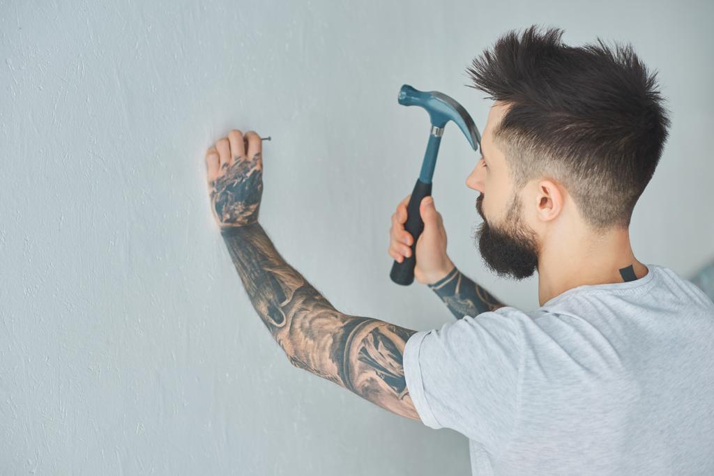 бородатий татуйований молодий чоловік забиває цвях на стіні
  - Фото, зображення