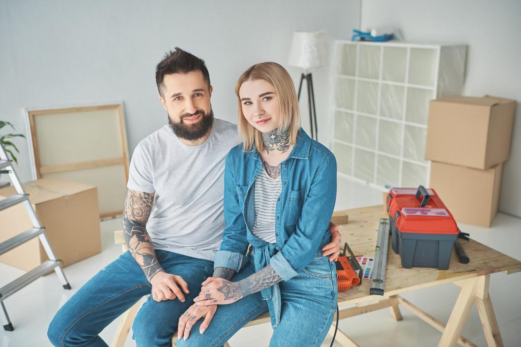 happy young couple in tattoos sitting together and smiling at camera while making repairs in new house - Photo, Image
