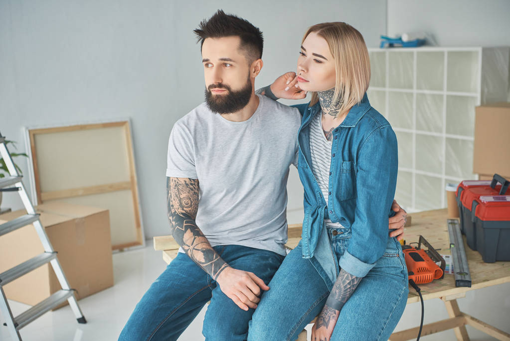 jeune couple avec des tatouages assis ensemble et regardant loin dans une nouvelle maison
 - Photo, image