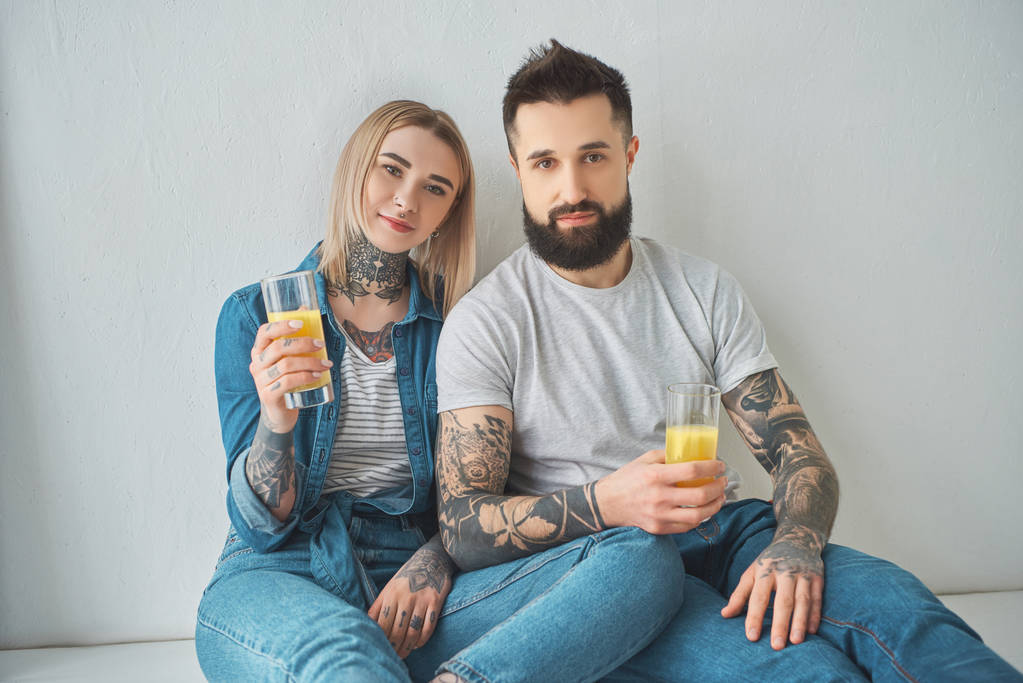 νεαρό ζευγάρι τατουάζ κρατώντας ποτήρια χυμό και βλέπουν φωτογραφική μηχανή, ενώ κάθεται στο πάτωμα στο καινούργιο σπίτι - Φωτογραφία, εικόνα