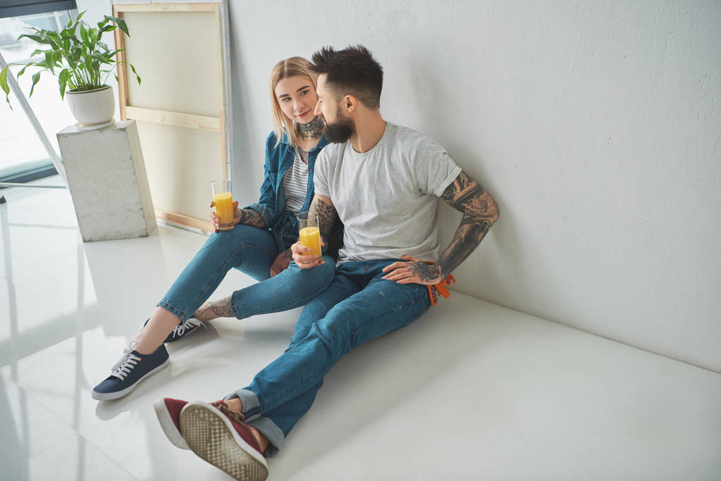 feliz jovem tatuado casal segurando copos de suco e sentado no chão em nova casa
  - Foto, Imagem