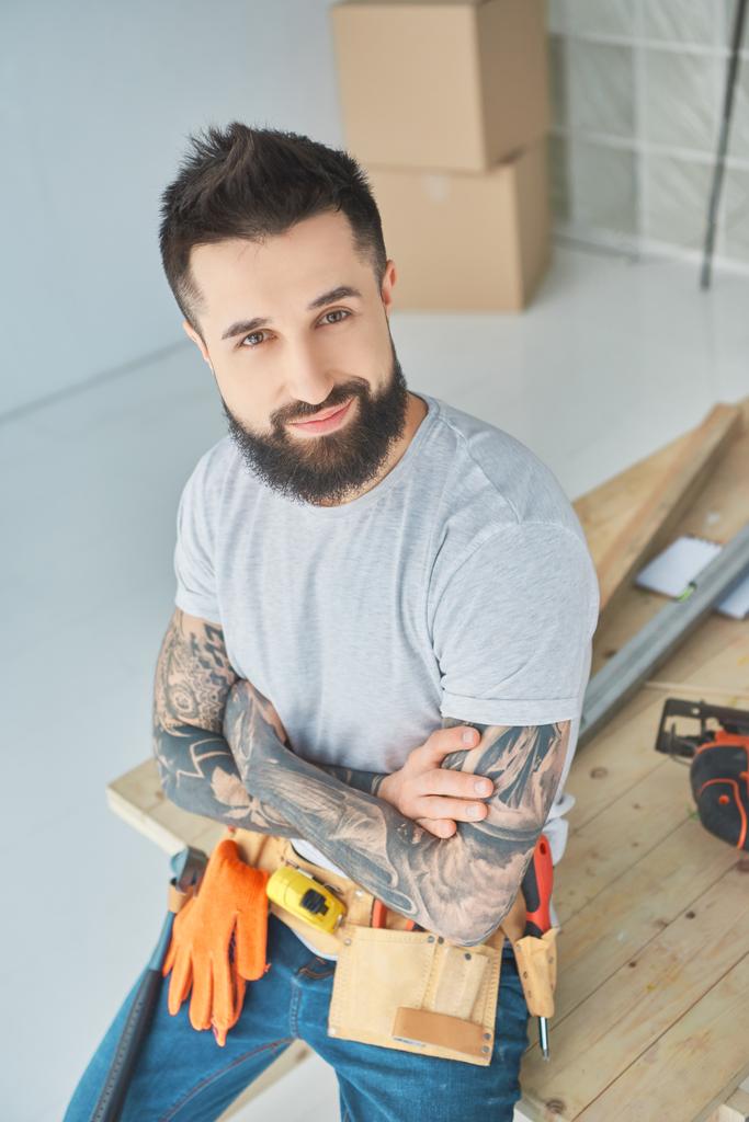 reparador sonriente con tatuajes y herramientas apoyadas en la superficie de madera en el nuevo apartamento
 - Foto, Imagen