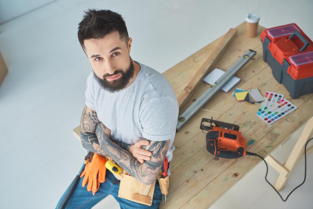 high angle view of bearded repairman with tattoos leaning on wooden table with equipment - Photo, Image